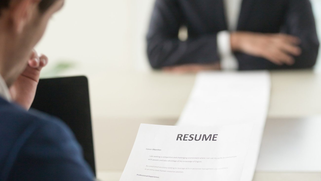 Back view of employer reading resume focus on document, job applicant waiting for hiring decision at desk on background. Hiring manager studying long list of candidates achievements and experiences